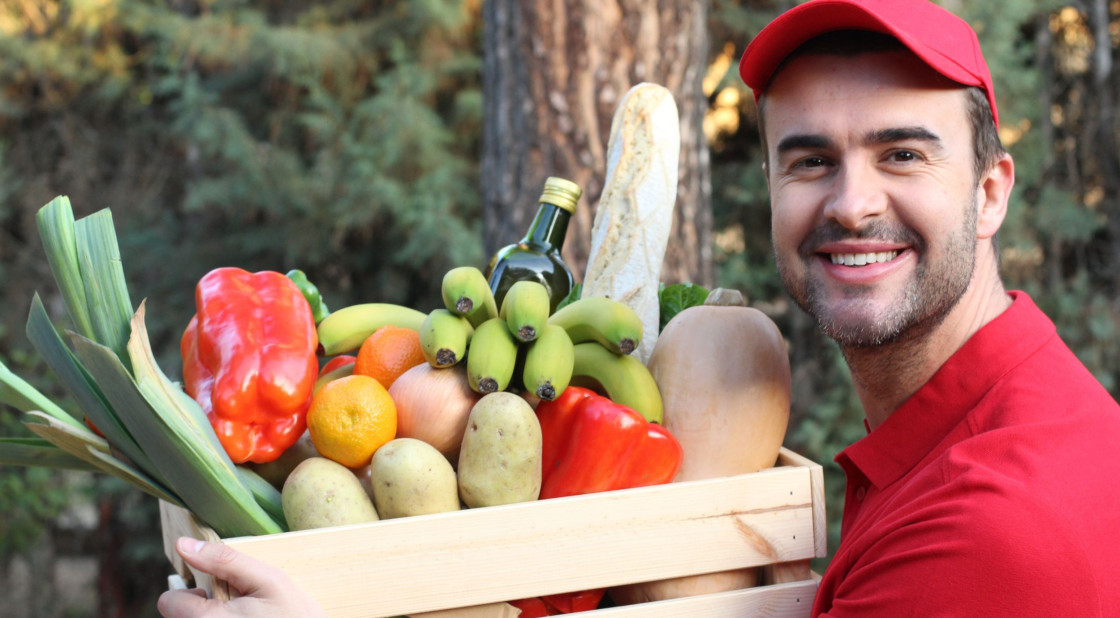 Cheerful man holding a crate of fresh fruits and vegetables in a vibrant outdoor setting.