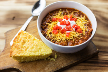 Red Chili In A Small White Bowl With A Piece Of Cornbread. Wood Background With Copy Space.